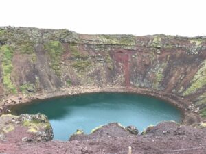 A 10-day Itinerary to Iceland's Ring Road 3 A bright blue-green water sits in the crater of Grabok Crater. The soil is red and the sides of the crater have moss and green grass leading down the sides.
