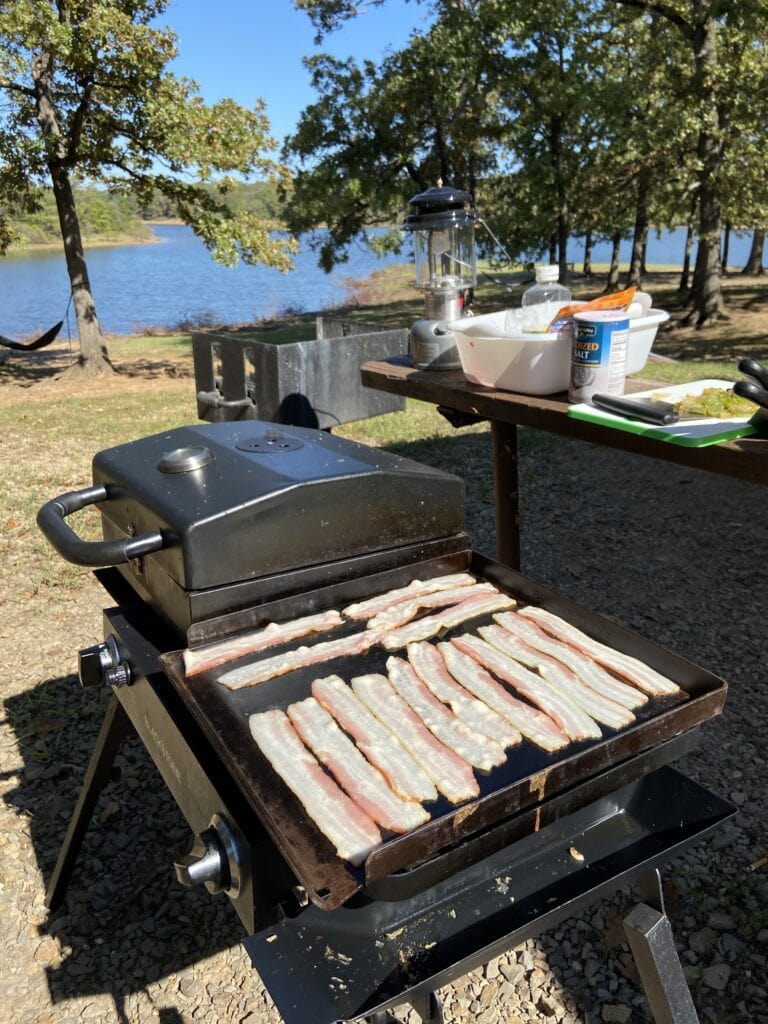 Easy Campfire Dinner Recipes for Families 6 Bacon cooking on a Blackstone Tailgator. A lake is in the distance and trees in front of the lake.