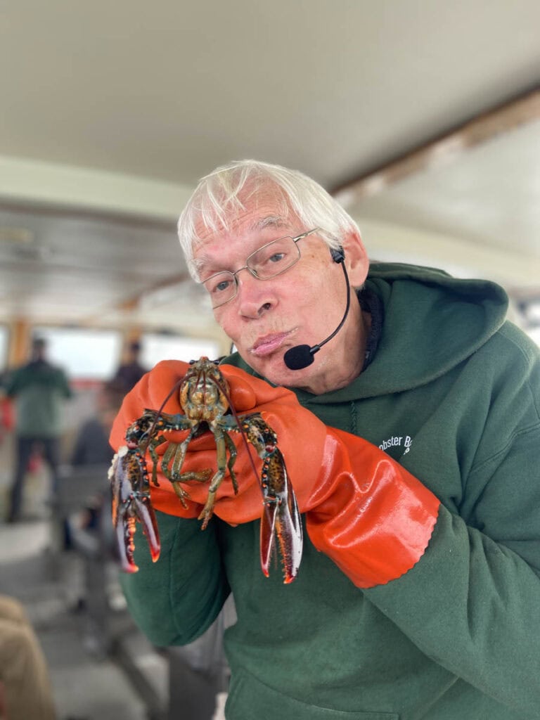 A tour guide with white hair and a microphone shows a pouty face, mimicing the face of the lobster that he is holding up in front of him.
