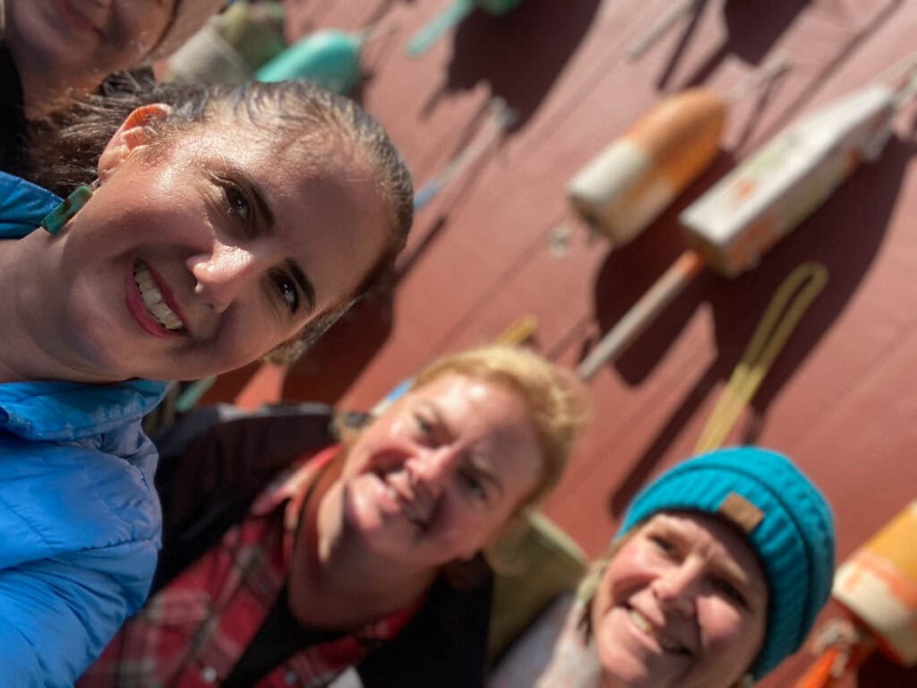 The author and two friends stand in front of a red building that is blurred, with lobster buoys adorning the walls.