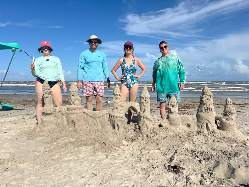 You should Be Learning How to Build Sand Castles in Port Aransas 1 4 people stand behind a large sand castle with blue sky in the background.